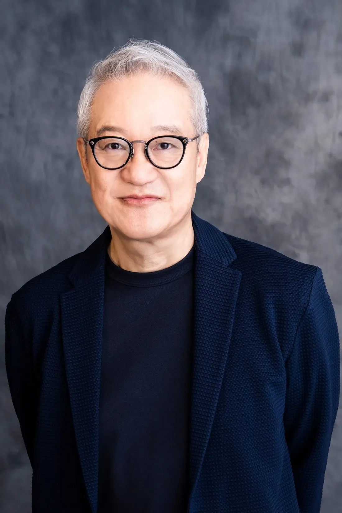 Headshot portrait of a man with short, graying hair wearing round, dark-framed glasses. He has a calm expression and is wearing a dark navy blue textured blazer over a dark navy blue t-shirt. The background is a mottled dark gray, providing a neutral backdrop that emphasizes the subject. The image style is a professional headshot, likely for a website or publicity purposes.