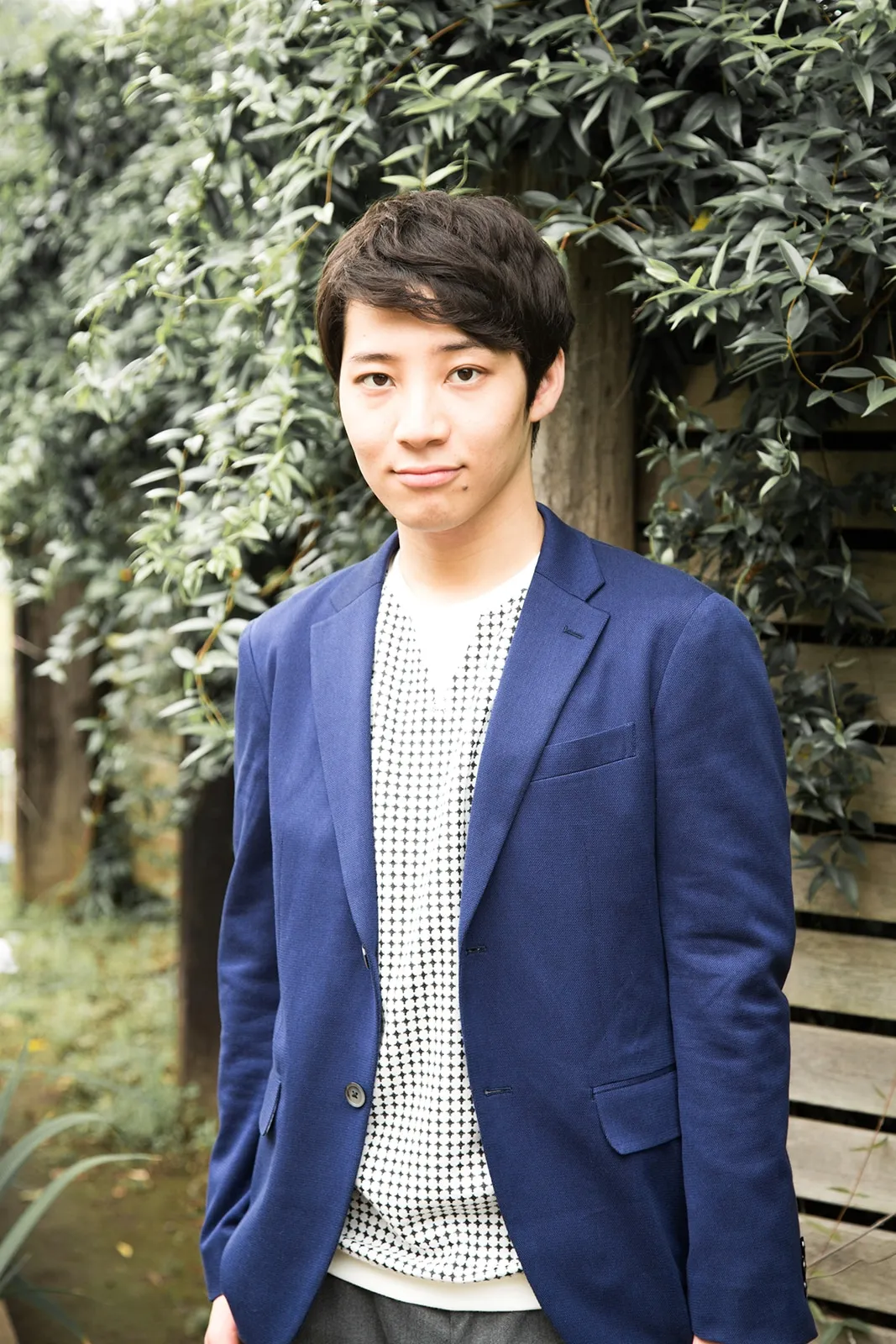 A young Asian man with short, dark brown hair stands slightly angled to the left, looking directly at the camera with a calm expression. He's wearing a dark blue blazer over a white shirt with a small, repeating black geometric pattern. The shirt is visible at the collar and beneath the blazer's open front. His pants appear to be dark gray. The background is softly blurred, showing a wall partially obscured by lush green foliage with small, oval leaves. The overall style of the image is a clean, simple portrait with a slightly natural feel.
